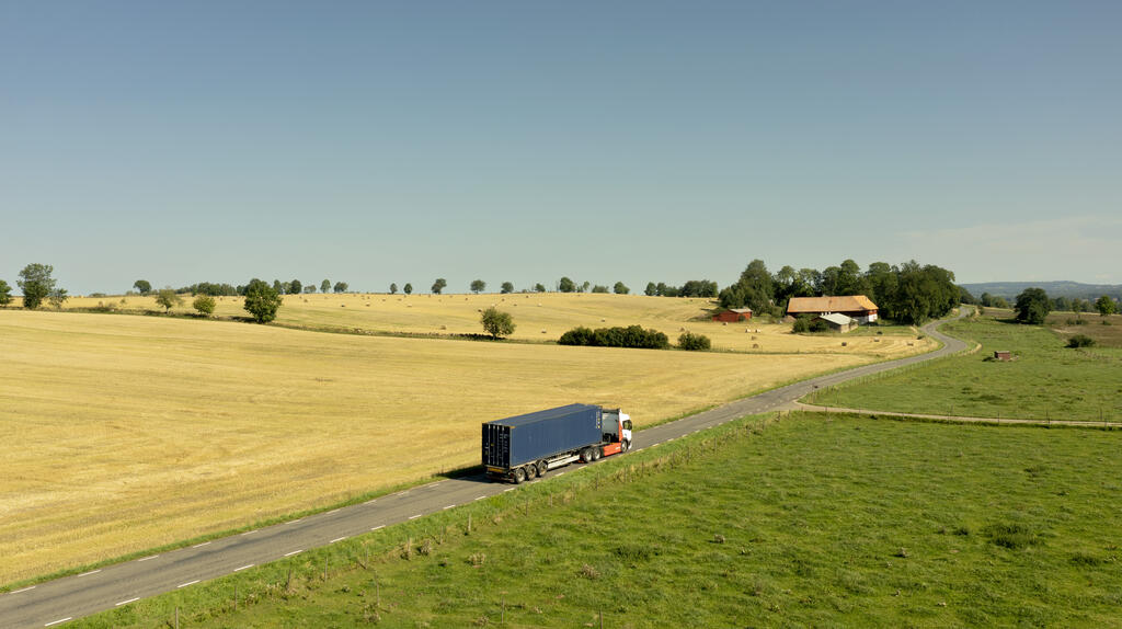Lastbil åker genom ett soligt åkerlandsskap på en landsväg. Solen skiner och grödorna är i högform. Somrig och harmonisk känsla speglar bilden.  