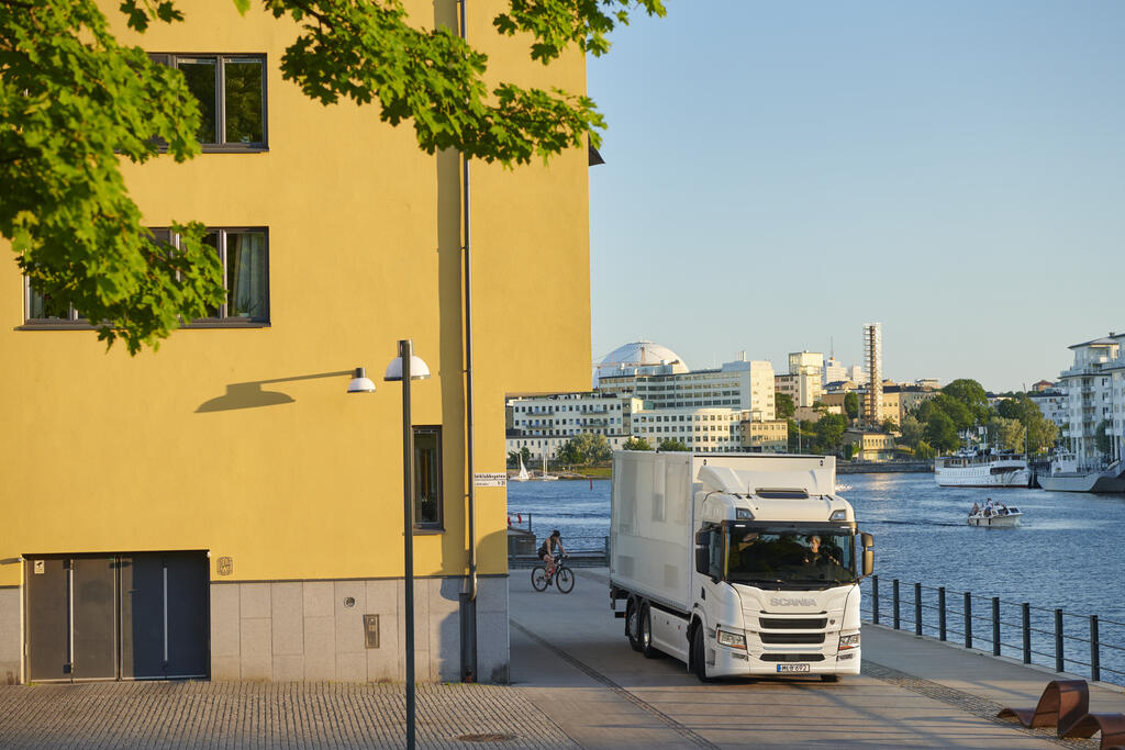 Lastbild som kör i stadsmiljö i kvällssolen. Ett fint hamninlopp öppnar upp sig i bakgrunden av lastbilen och stadsmiljön.